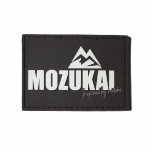 Peut inclure: Patch noir avec des coutures blanches autour des bords. Le patch présente un graphique de chaîne de montagnes blanches et le texte "MOZUKAI Inspiré par la nature".