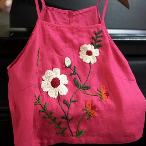 May include: A hot pink camisole top with embroidered floral details. The top features white flowers with red centers, orange flowers, and green leaves. The top is hanging on a wooden hanger.