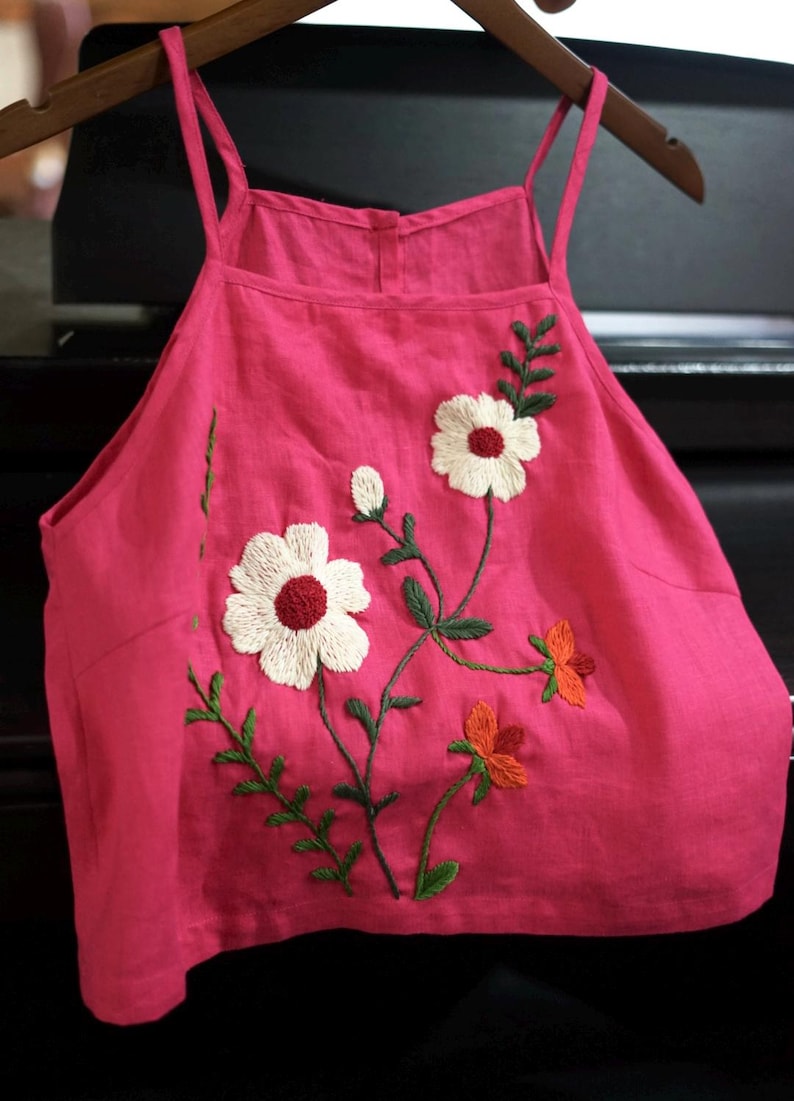 May include: A hot pink camisole top with embroidered floral details. The top features white flowers with red centers, orange flowers, and green leaves. The top is hanging on a wooden hanger.