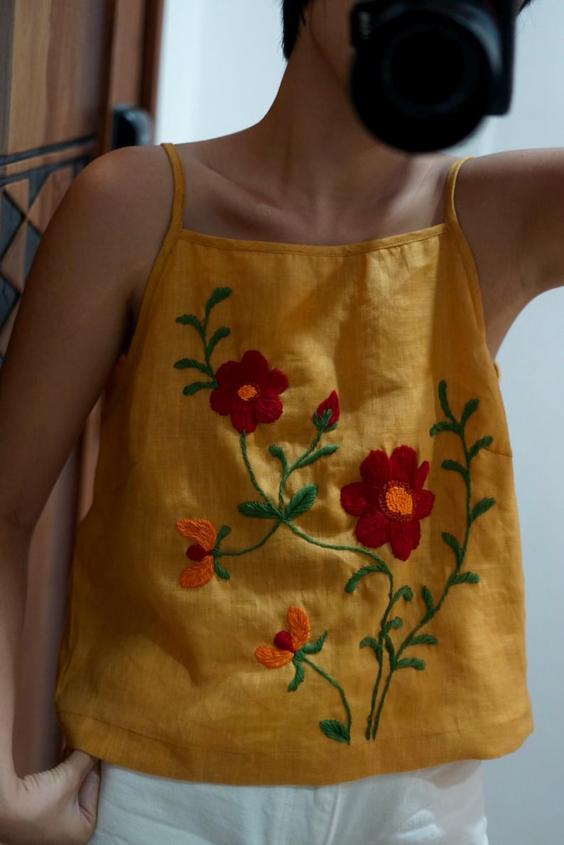 May include: Mustard yellow tank top with embroidered floral design. The top features red and orange flowers with green stems and leaves. The tank top has thin straps and a square neckline. The garment is made of a soft, textured fabric.