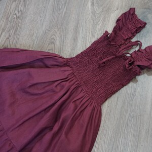 May include: A burgundy off-the-shoulder dress with a smocked bodice and ruffled sleeves. The dress features a flowing skirt and tie details at the neckline. The fabric appears to be a lightweight material, suitable for warm weather.