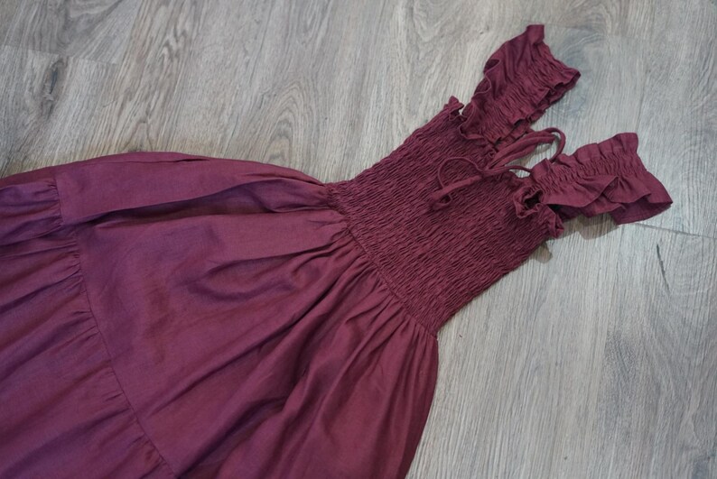 May include: A burgundy off-the-shoulder dress with a smocked bodice and ruffled sleeves. The dress features a flowing skirt and tie details at the neckline. The fabric appears to be a lightweight material, suitable for warm weather.