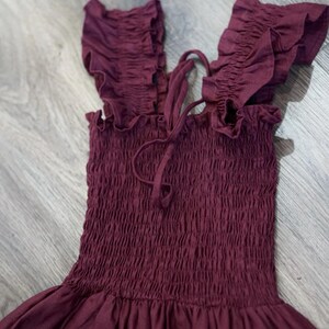 May include: A burgundy-colored dress with a smocked bodice and ruffled shoulder straps. The dress has a flowing skirt and a tie detail at the neckline. The fabric appears to be a soft, lightweight material, suitable for warm weather.