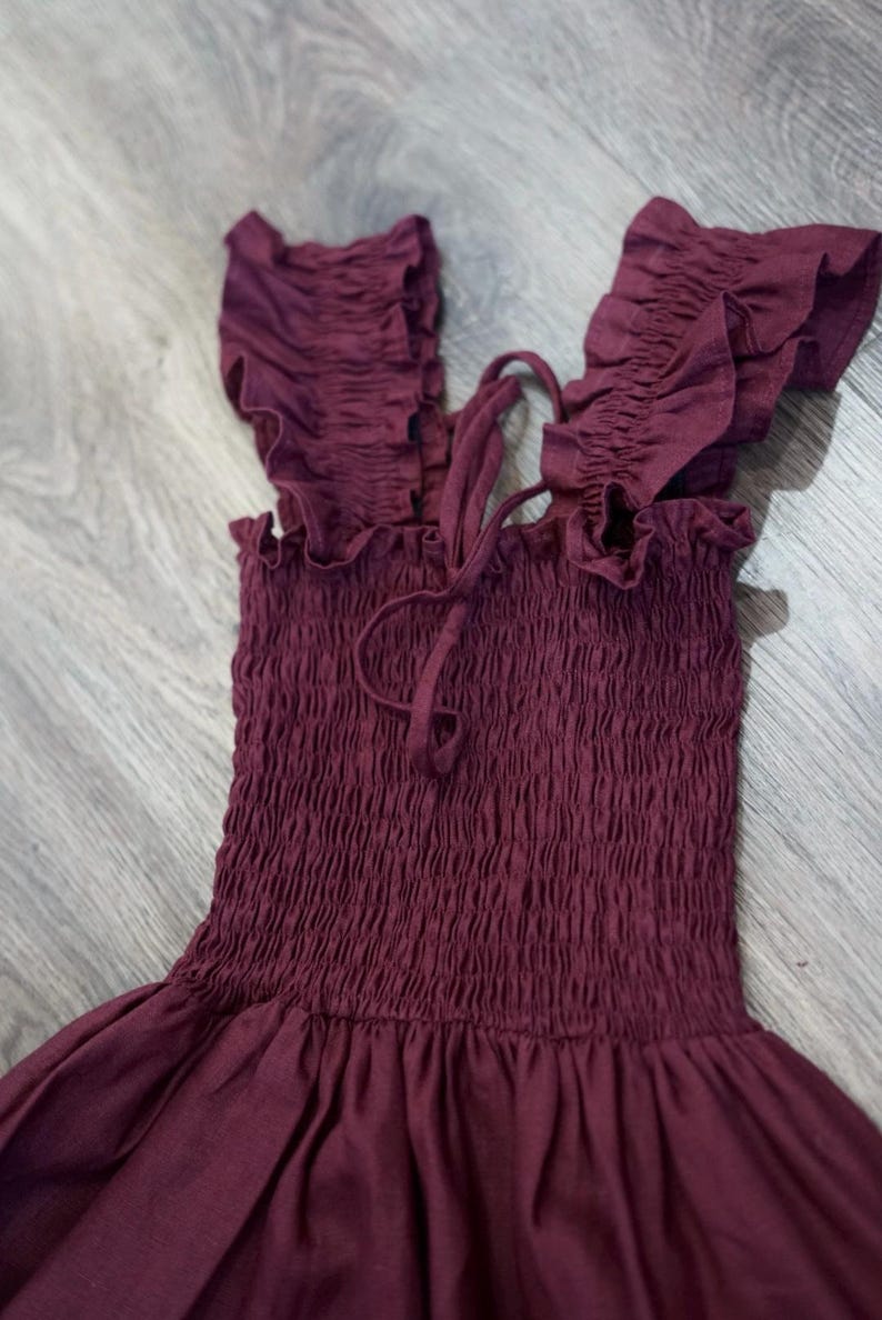 May include: A burgundy-colored dress with a smocked bodice and ruffled shoulder straps. The dress has a flowing skirt and a tie detail at the neckline. The fabric appears to be a soft, lightweight material, suitable for warm weather.
