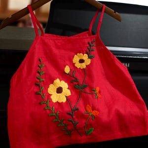 May include: A red camisole top with yellow and orange floral embroidery. The top has thin straps and is hanging on a wooden hanger. The embroidered flowers have yellow petals and brown centers, with green stems and leaves.