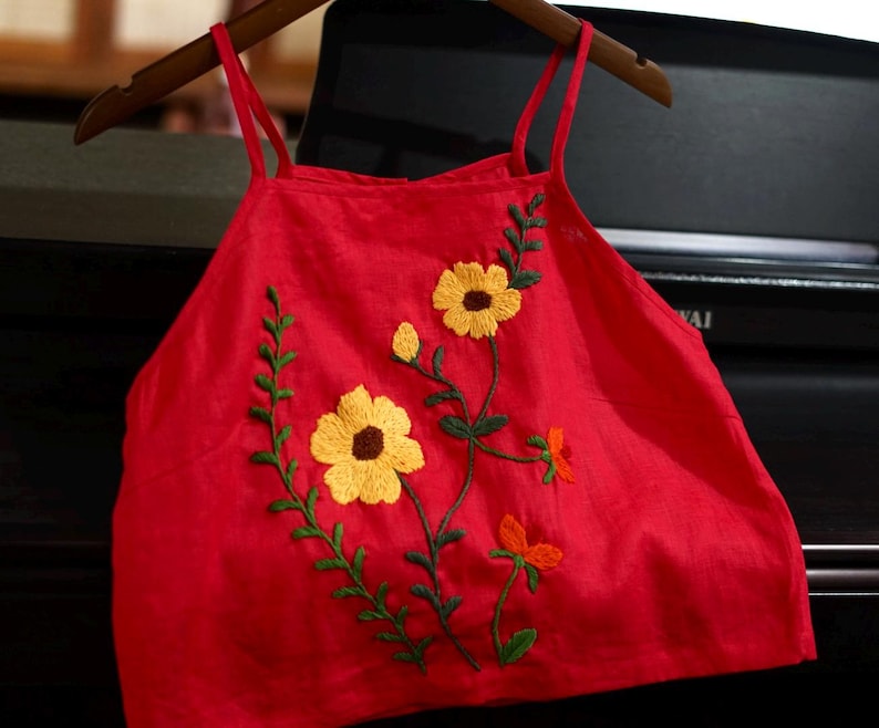 May include: A red camisole top with yellow and orange floral embroidery. The top has thin straps and is hanging on a wooden hanger. The embroidered flowers have yellow petals and brown centers, with green stems and leaves.