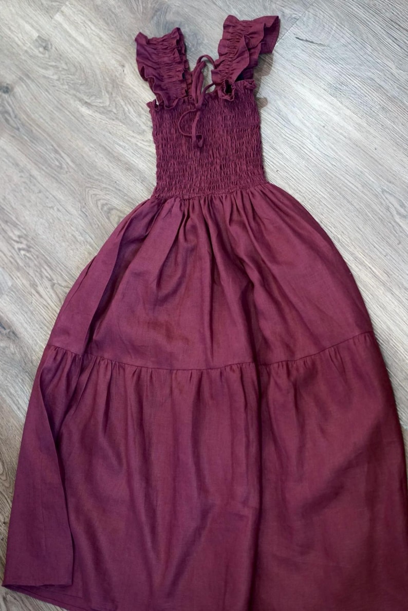 May include: A burgundy-colored dress with a smocked bodice and ruffled straps. The dress has a flowing, tiered skirt. The straps are tied at the top. The dress is made of a soft, lightweight fabric, perfect for warm weather.