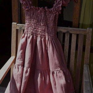 May include: A dusty rose-colored dress with a smocked bodice and tiered skirt hangs on a wooden hanger. The dress features ruffled cap sleeves and a drawstring neckline. The dress is made of a lightweight fabric and is displayed on a wooden chair.