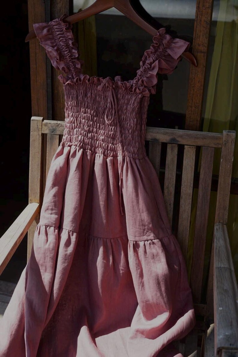 May include: A dusty rose-colored dress with a smocked bodice and tiered skirt hangs on a wooden hanger. The dress features ruffled cap sleeves and a drawstring neckline. The dress is made of a lightweight fabric and is displayed on a wooden chair.