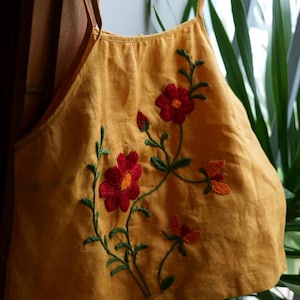 May include: Mustard yellow top with embroidered red flowers and green stems. The top has thin straps and a scoop neckline. The floral embroidery adds a decorative touch to the garment. The top is hanging on a wooden hanger.