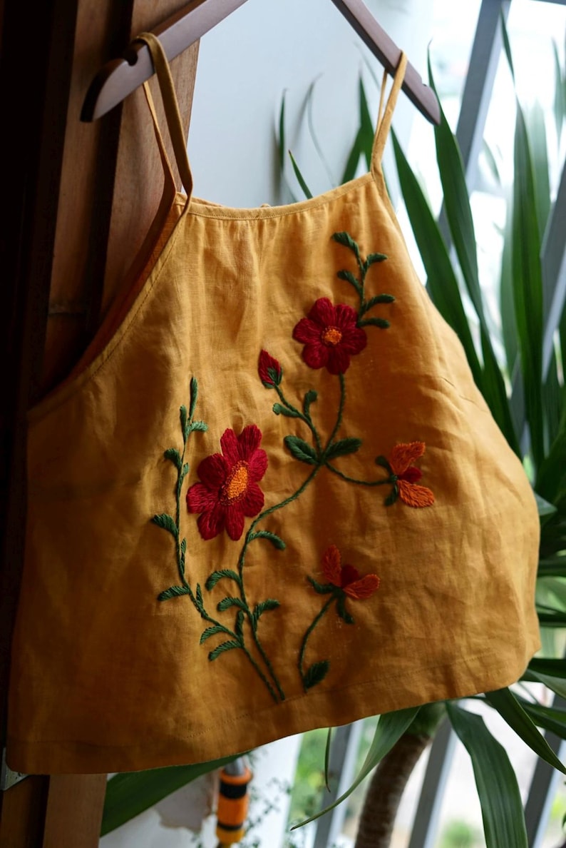 May include: Mustard yellow top with embroidered red flowers and green stems. The top has thin straps and a scoop neckline. The floral embroidery adds a decorative touch to the garment. The top is hanging on a wooden hanger.