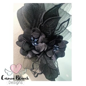 May include: Black floral hair comb with a delicate black mesh and blue jewel accents. The comb is adorned with two large black fabric flowers and a smaller black flower with a blue jewel center.