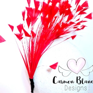 May include: A red feather spray with a heart-shaped design. The feathers are arranged in a radial pattern, creating a burst of color. The spray is attached to a black stem. The image also includes the text "Carmen Blanch Designs" with a heart and wings logo.