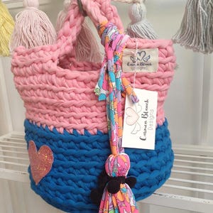 May include: Pink and blue crocheted basket with a heart-shaped design and a pink tassel. The basket has a tag that says "Carmen Blanch Designs".