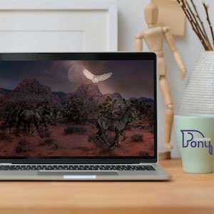 May include: A laptop computer displaying a digital image of a desert landscape with a full moon, a white owl flying, and a group of black horses running. The laptop is on a wooden table with a green mug and a vase with branches.