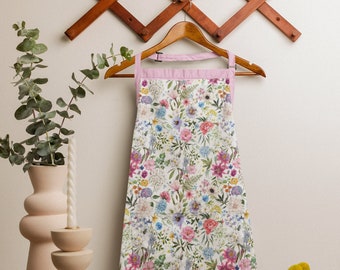 May include: A white apron with a floral print in shades of pink, blue, and yellow. The apron has a pink trim and a pocket on the front.