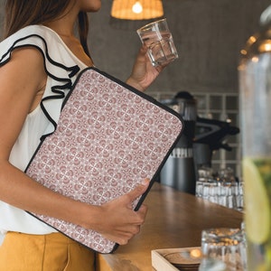 Peut inclure: Une femme tenant une housse d'ordinateur portable avec un motif géométrique rose et blanc. La housse a une fermeture éclair noire et une bordure noire.