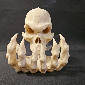 Puede incluir: Un portavelas con forma de calavera beige con una expresión amenazante. La calavera está sujeta por dos manos esqueléticas. El portavelas está hecho de un material de color claro y se asienta sobre una superficie oscura. Un artículo decorativo único.