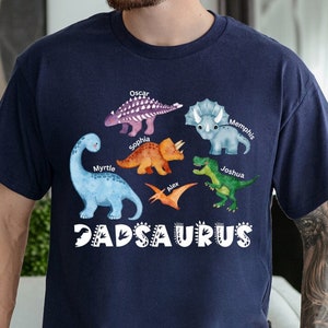May include: A navy blue t-shirt featuring a graphic of colourful dinosaurs and the text "DADSaurus". The dinosaurs are labelled with names such as "Oscar", "Sophia", "Myrtle", "Memphis", "Joshua", and "Nick".