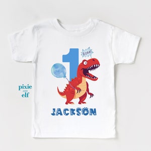 以下が含まれることがあります： 赤い恐竜が青い風船を持っている白い子供用Tシャツ。大きな青い数字の1が恐竜の隣にあり、「ROAR！」という言葉が吹き出しの中にあります。名前の「JACKSON」は下に青色で印刷されています。