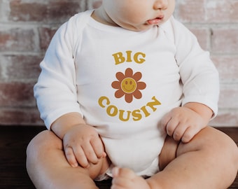 May include: A white long-sleeved onesie with the text "BIG COUSIN" in yellow letters with a brown flower with a yellow smiley face in the center.