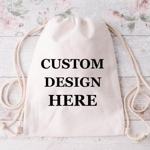 May include: A cream-coloured drawstring bag with the words "CUSTOM DESIGN HERE" printed in black. The bag has a simple design with rope-like drawstrings.
