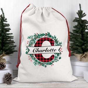 Peut inclure: Un sac cadeau de Noël blanc avec un cordon de serrage rouge. Le sac présente un motif de couronne avec la lettre "C" à carreaux rouges et noirs et le nom "Charlotte" sur une bannière blanche. Deux petits sapins et des pommes de pin encadrent le sac.