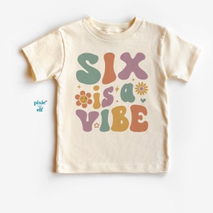 Pode incluir: Uma camiseta bege claro com o texto "SIX is a VIBE" em letras retrô coloridas. O texto é cercado por flores e estrelas.