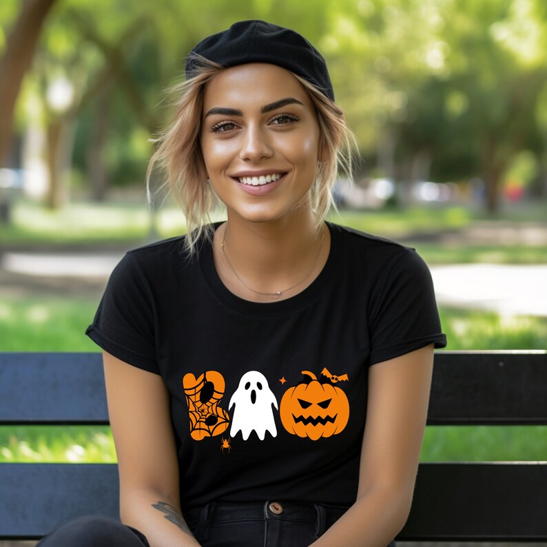K&ouml;nnte beinhalten: Ein schwarzes T-Shirt mit einer orangefarbenen Grafik eines Geistes, eines Spinnennetzes, einer Spinne, einer Fledermaus und eines K&uuml;rbisses mit dem Wort "BOO" in Orange.