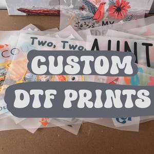 May include: A selection of DTF (Direct to Film) prints, showcasing diverse designs and text. The prints include images of a construction vehicle, floral arrangements, and phrases like "Two, Two" and "Auntie". The words "CUSTOM DTF PRINTS" are displayed.