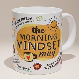 May include: White ceramic mug featuring the text "the MORNING MINDSET mug" in black and orange. The mug is decorated with a yellow sun, a coffee cup with a heart, and other motivational phrases. It has a white handle.