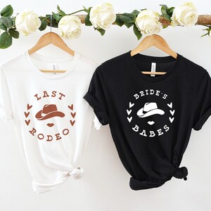 May include: Two white t-shirts with a brown cowboy hat graphic and the text "Last Rodeo" and "Bride's Babes" printed on them.