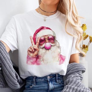 May include: White t-shirt featuring a pink and white illustration of Santa Claus wearing sunglasses and blowing a bubble. Santa is giving the peace sign.