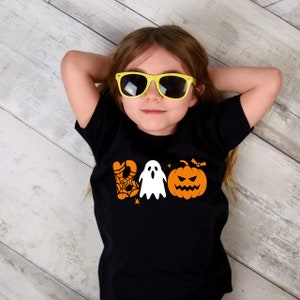 K&ouml;nnte beinhalten: Ein Kind tr&auml;gt ein schwarzes T-Shirt mit einer orangefarbenen und wei&szlig;en "BOO"-Grafik mit einem Geist, einem K&uuml;rbis, einem Spinnennetz und Flederm&auml;usen. Das Kind tr&auml;gt eine gelbe Sonnenbrille und hat die Arme hinter dem Kopf.