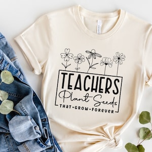 May include: A beige t-shirt with a black graphic that reads "Teachers Plant Seeds That Grow Forever". The graphic features a row of five stylised flowers.