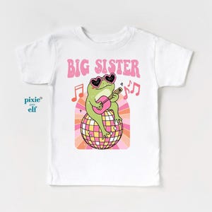 May include: White t-shirt featuring a pink and green graphic of a frog wearing sunglasses and playing a guitar on a disco ball. The text "BIG SISTER" is above the frog.