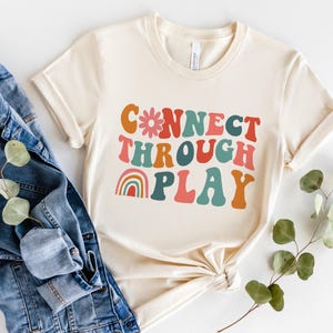 May include: A cream-coloured t-shirt featuring the phrase "CONNECT THROUGH PLAY" in a retro, colourful font, with a flower and rainbow graphic. The shirt is styled with a denim jacket and sprigs of greenery.