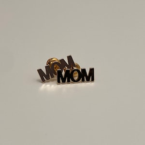 Op de afbeelding: Drie gouden pinnen met het woord "MOM" in zwarte letters. De pinnen zijn op elkaar gestapeld.