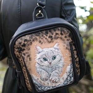 以下が含まれることがあります： 黒の革製のバックパックで、茶色と黒のヒョウ柄のデザインです。バックパックの前面には、青い目をした白い子猫の手描き画像が描かれています。