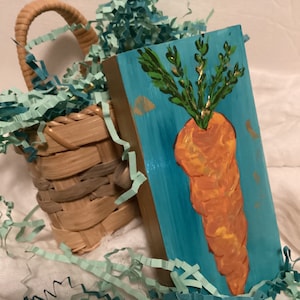 May include: A hand-painted wooden block with a bright orange carrot and green leafy top on a turquoise background. The carrot is painted in a stylized way with orange, yellow, and brown hues. The block is surrounded by green and blue tissue paper.