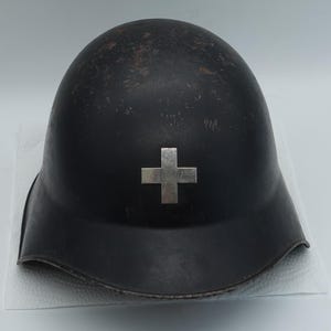 Può includere: Un elmetto in metallo grigio antracite scuro con un emblema a croce color argento centrato sulla parte anteriore. L'elmetto presenta una forma a cupola liscia e arrotondata con una tesa leggermente svasata e segni di usura superficiale.