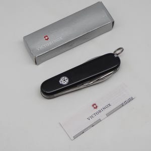 Könnte beinhalten: Ein schwarzes Schweizer Taschenmesser mit silbernem Schlüsselring, kleinem weißem Emblem und mehreren Werkzeugen. Das Messer liegt neben einer silbernen Schachtel und einem kleinen weißen Papier mit dem Markennamen.