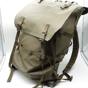 Könnte beinhalten: Ein olivgrüner Rucksack mit einer großen Deckelklappe, die mit zwei Kunststoff-Schnallenriemen gesichert ist. Der Rucksack verfügt über verstellbare Nylongurte, seitliche Kompressionsriemen und gepolsterte Schultergurte, alles in Olivgrün.