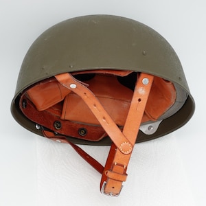 May include: An olive drab military helmet with a brown leather interior and chin strap. The helmet has a rounded shape and is designed to protect the head. The leather straps are secured with metal rivets and buckles.