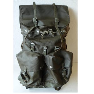 Puede incluir: Una mochila de lona color verde oliva con una solapa superior grande asegurada con dos correas de hebilla ajustables, un cierre de cordón con ojales metálicos y dos bolsillos delanteros espaciosos con hebillas y herrajes.