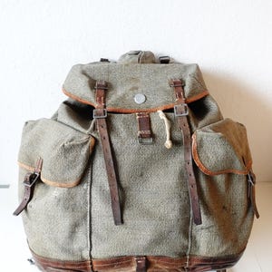 Könnte beinhalten: Vintage olivgrüner Rucksack mit braunen Lederriemen und Akzenten. Er verfügt über zwei Seitentaschen und einen oberen Klappenverschluss. Der Rucksack hat einen strukturierten Stoff und einen Metallverschluss. Der Boden des Rucksacks ist ebenfalls mit braunem Leder verkleidet.