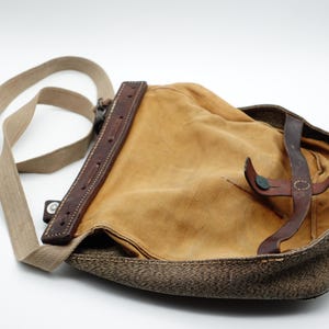 Puede incluir: Bolso de hombro de estilo vintage con cuerpo de lona color canela y correa y detalles de cuero marrón. El bolso tiene un ribete marrón tejido y una correa de hombro de lona beige. El bolso está abierto, revelando el interior.