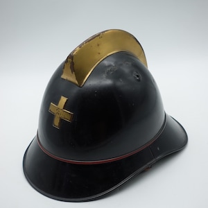 Può includere: Un casco nero con finitura lucida, caratterizzato da una prominente cresta color oro sulla parte superiore e da un emblema a croce color oro sul davanti. Una sottile linea rossa circonda la base del casco, dotato di tesa larga e cupola.