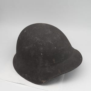 Può includere: Un elmetto da combattimento in metallo marrone scuro e usurato, con una cupola arrotondata e una tesa svasata. La superficie dell'elmetto mostra segni di usura, tra cui graffi, scalfitture e una finitura opaca e ruvida su tutta la calotta.