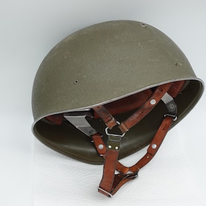 Può includere: Un elmetto militare verde oliva con una cupola liscia e arrotondata e un bordo in metallo. L'interno presenta un sistema di sospensione in pelle marrone con cinghie regolabili, fibbie in metallo e rivetti, offrendo una vista sulla struttura.